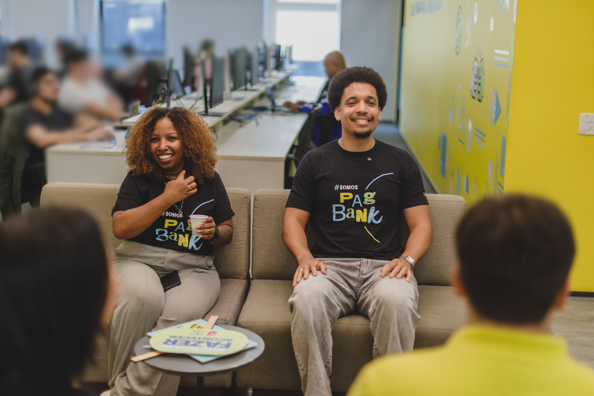 Dois PagTalents sorrindo no sofá do escritório PagBank com camisetas #SomosPagBank